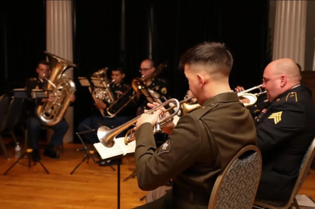 A trumpet player dressed in military attire performing with a large brass ensemble at a formal event.